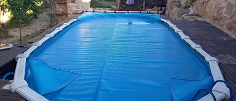 Une piscine extérieure recouverte d’une bâche bleue, entourée de murs en pierre et d’une terrasse en bois.