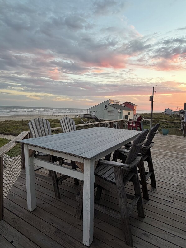 A wooden deck with a table and chairs, overlooking a beach with houses in the background.