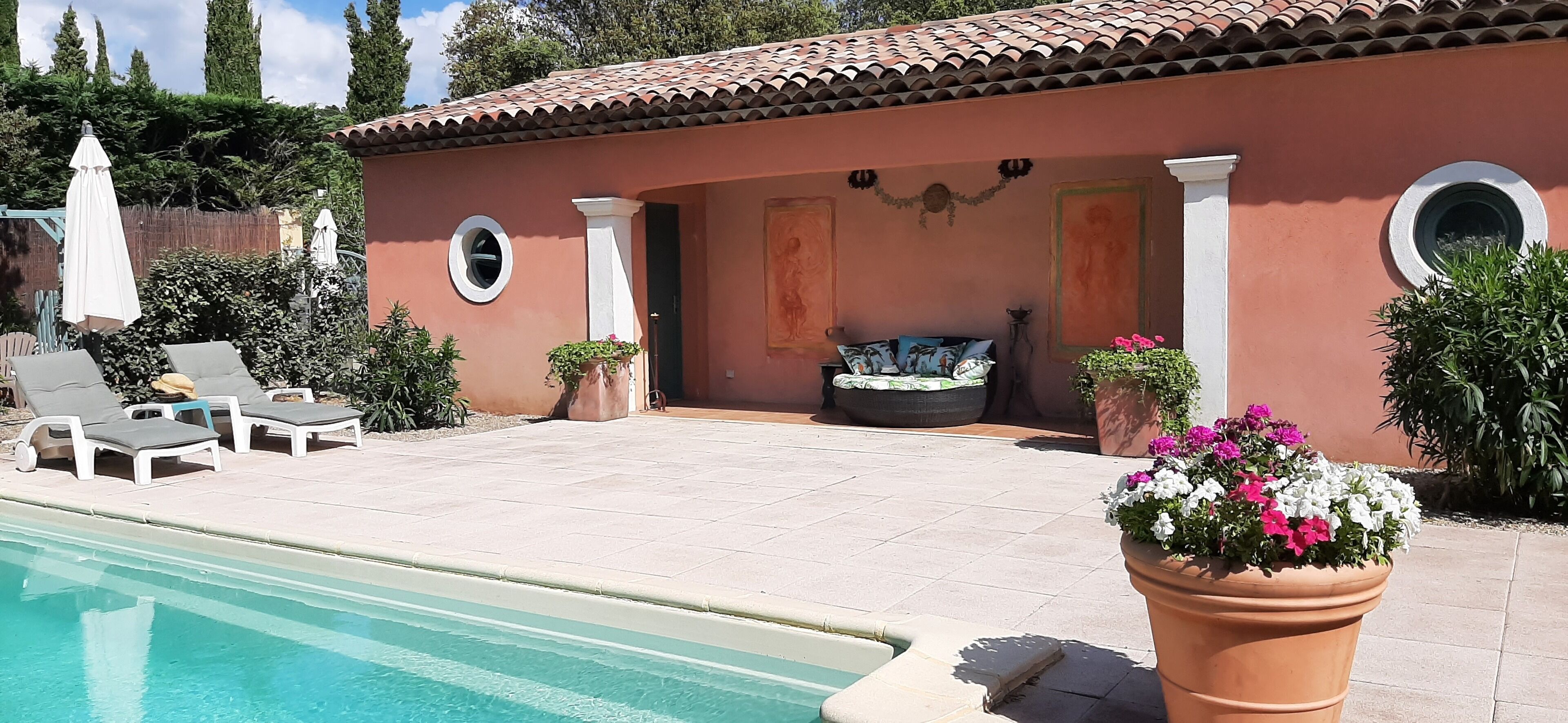 Un espace aménagé au bord de la piscine, avec des chaises longues, un petit coin salon doté de coussins et des plantes en pot.