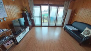 A spacious living room with wooden flooring, a flat-screen TV, a sofa, and a sliding glass door leading to an outdoor area.