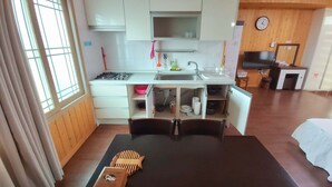 A compact kitchen with white cabinets, a stove, and a sink. There is a dining table with a black tablecloth and wooden blocks. A clock and a TV are visible in the background.