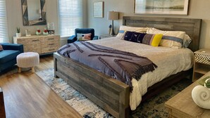 Un dormitorio con cama de madera, un sillón azul, un otoman redondito, un aparador y un espejo grande en la pared.