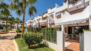 A modern hotel with white exterior walls, balconies with hanging plants, and a paved walkway with palm trees. - Apartamento Rosa (Mar de Pulpi) (San Juan de los Terreros)