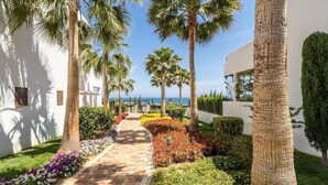 A pathway lined with palm trees and flowering plants leading to a building with a white facade. - Apartamento Rosa (Mar de Pulpi) (San Juan de los Terreros)