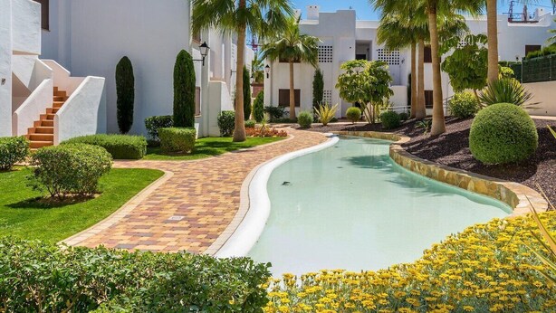 A pool surrounded by white buildings with palm trees and a brick pathway. - Apartamento Rosa (Mar de Pulpi) (San Juan de los Terreros)