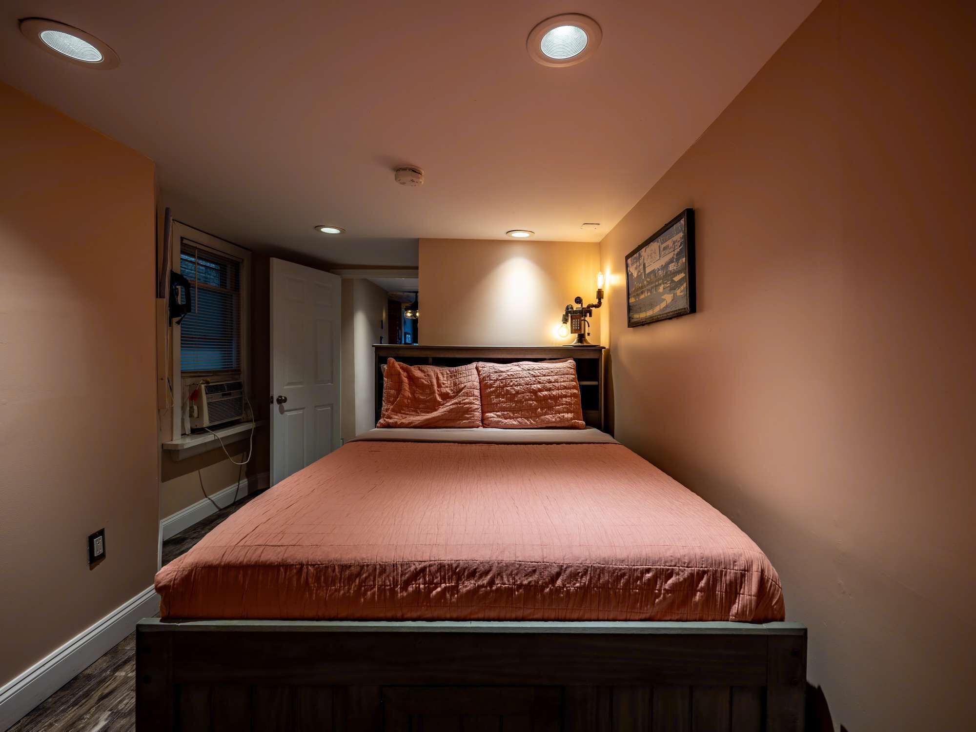 A bedroom with a bed, a nightstand, a wall-mounted lamp, and a window with blinds.