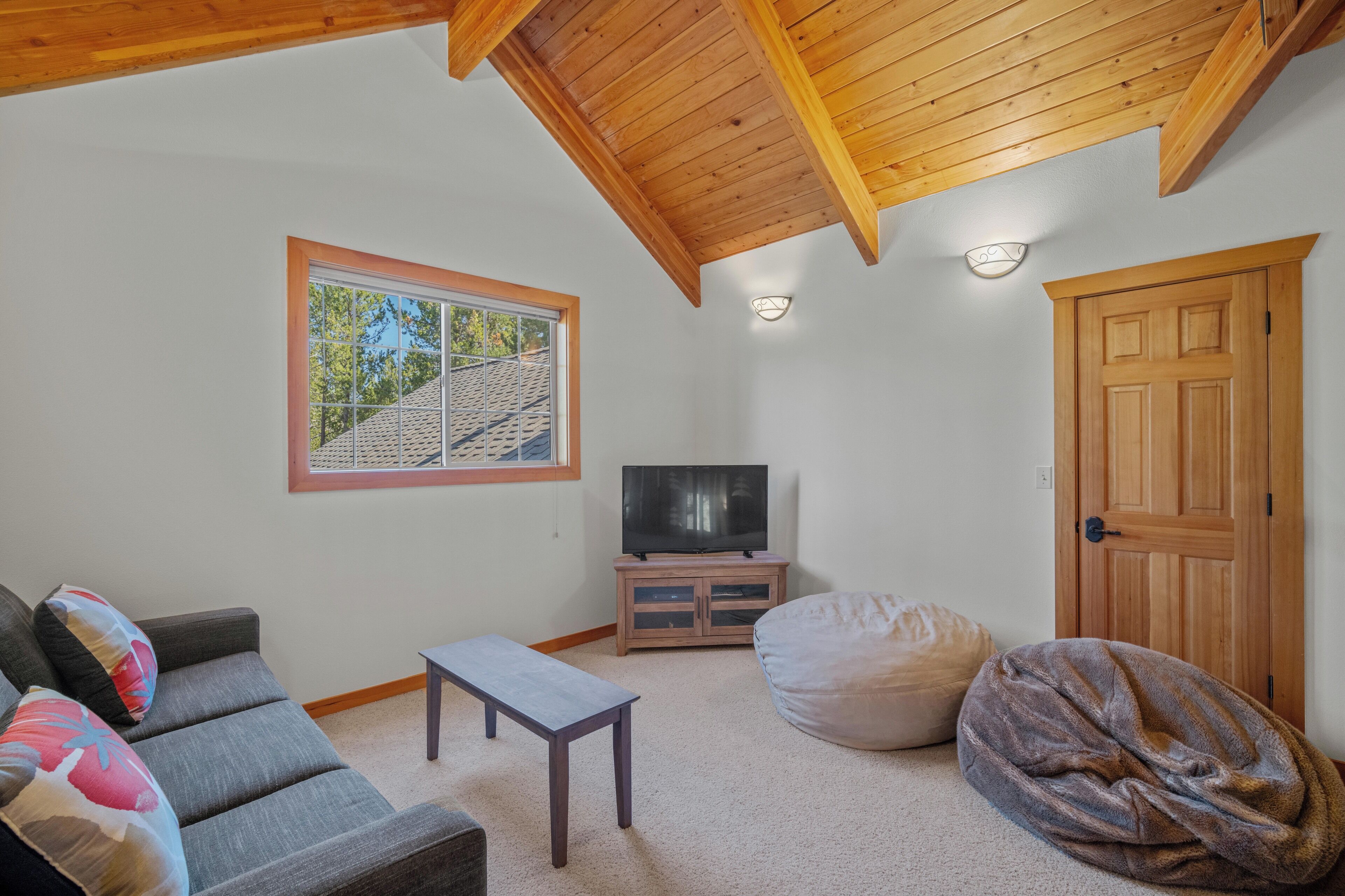A cozy living room with a grey sofa, a small table, a bean bag, a TV on a wooden cabinet, and a large window with a view of a house.