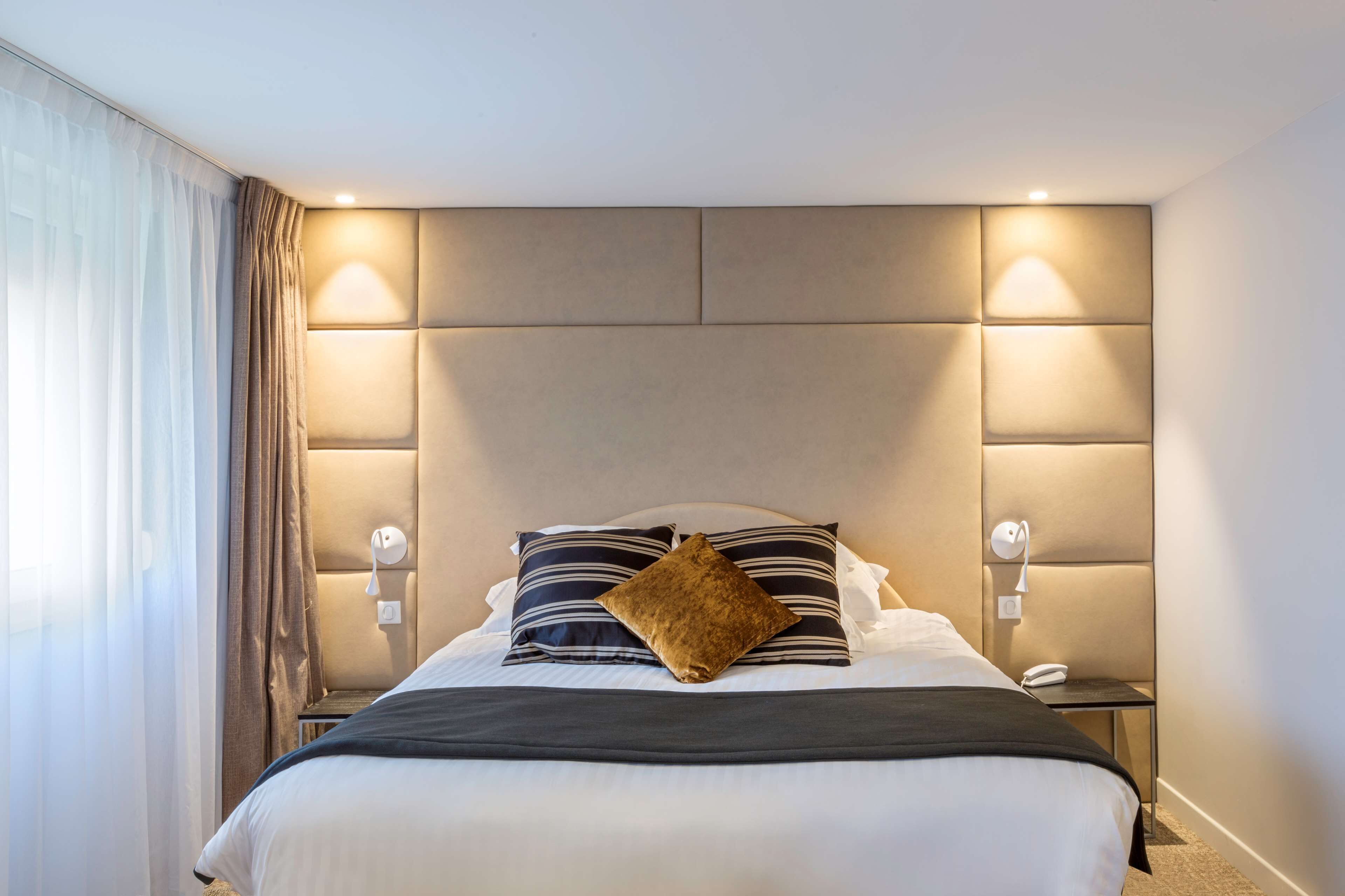 Una habitación de hotel con una cama grande, cabecera con iluminación integrada, mesita de noche con teléfono y ventana con cortinas.
