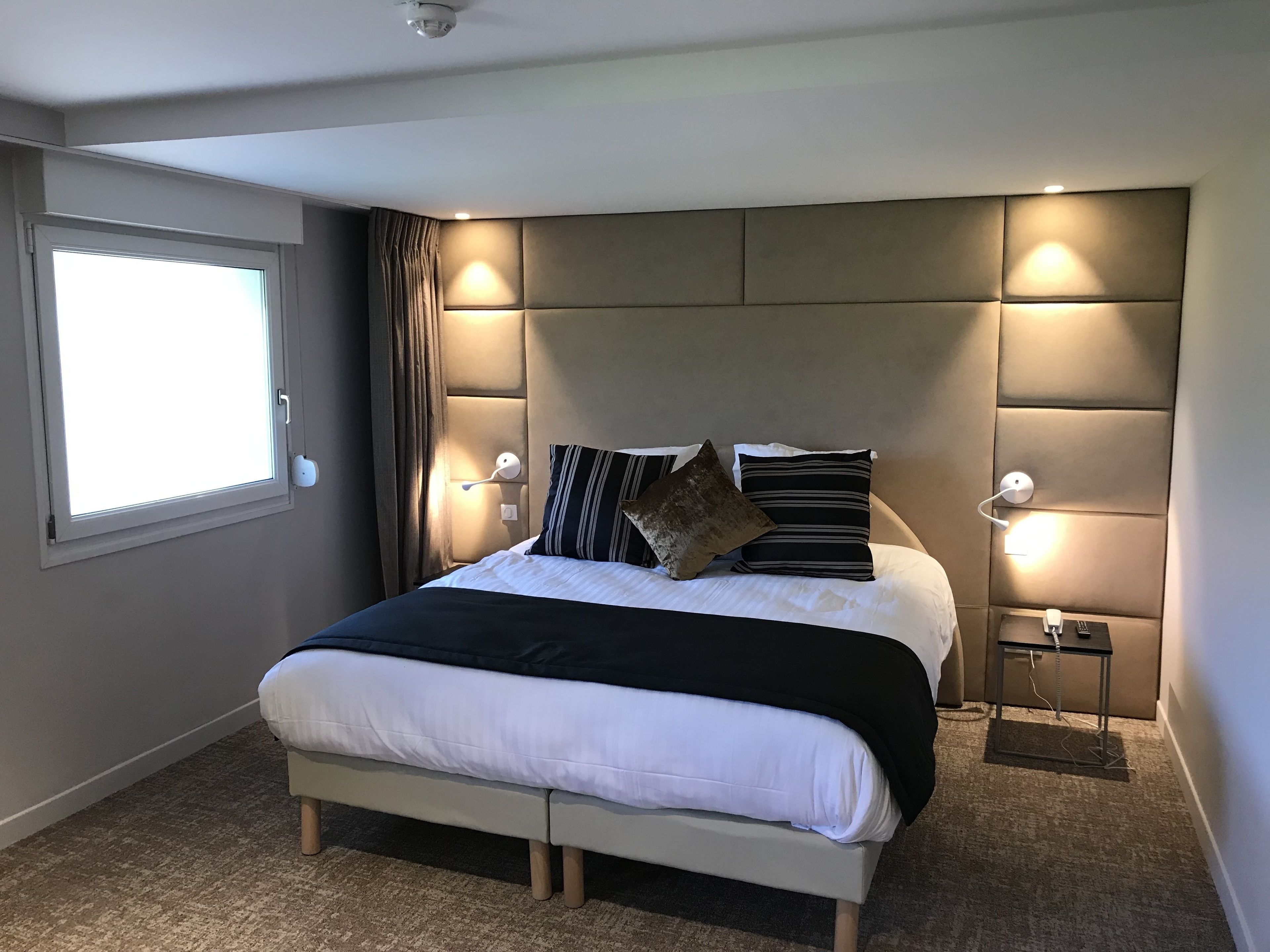 Een moderne hotelkamer met een groot bed, nachtlampjes, een hoofdeinde met ingebouwde verlichting, een raam met gordijnen, en een klein nachtkastje met een telefoon.
