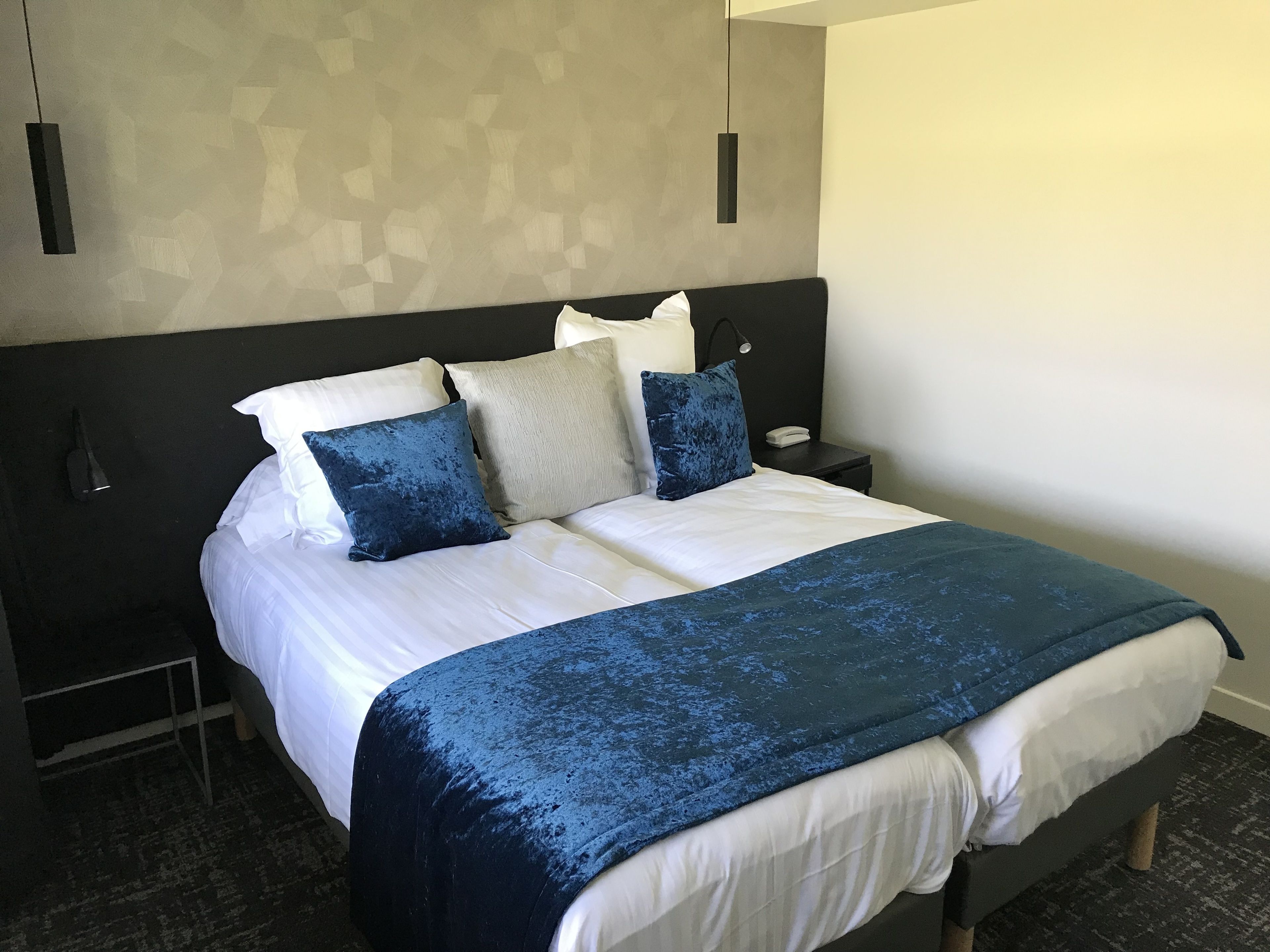 A neatly made bed with a white and blue bedspread, a dark headboard, and multiple pillows in shades of white, blue, and grey.