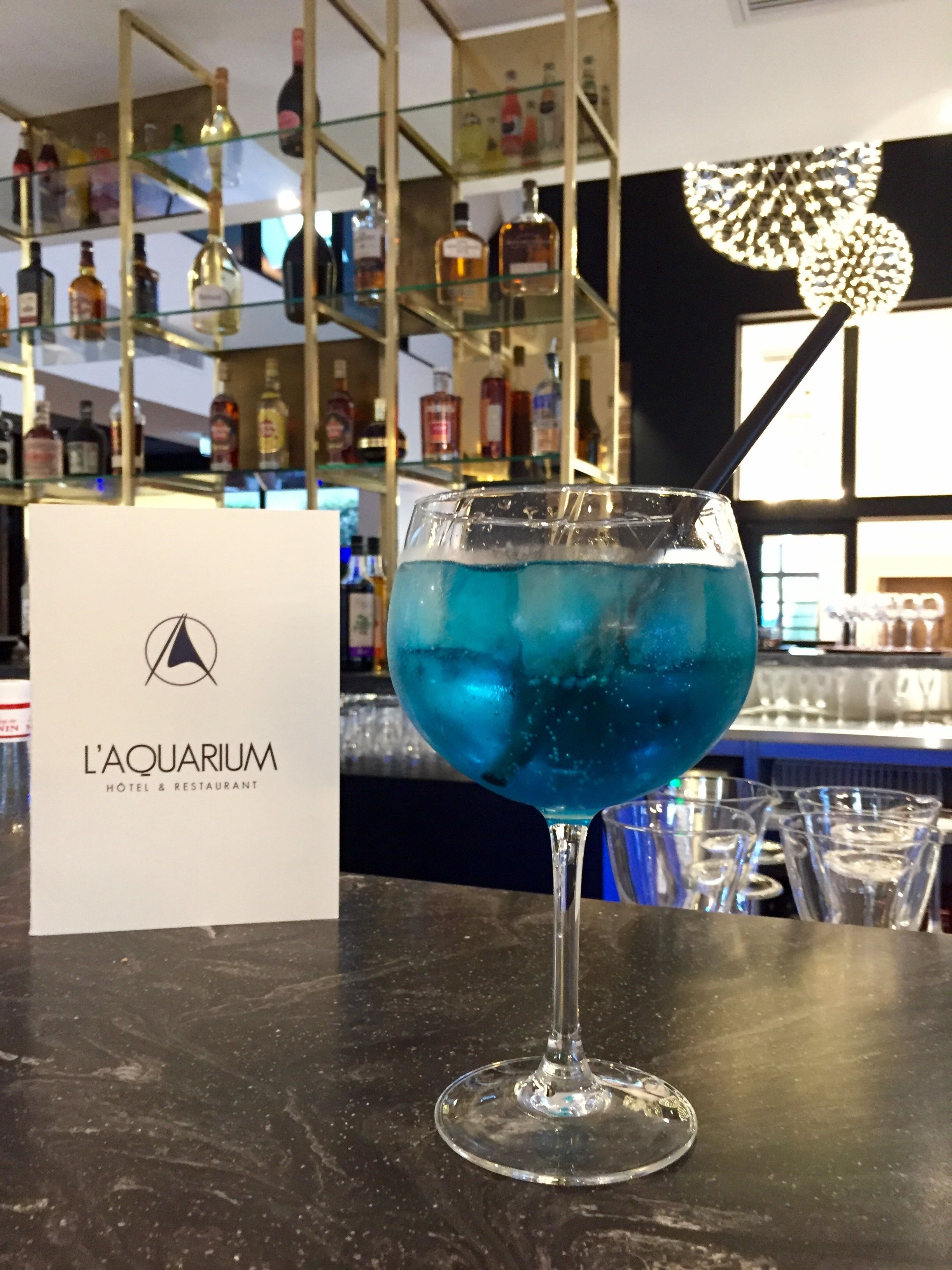 Een cocktailglas met een blauwe drank op een barkruk, een bordje voor L'AQUARIUM HOTEL & RESTAURANT, en planken met diverse flessen op de achtergrond.