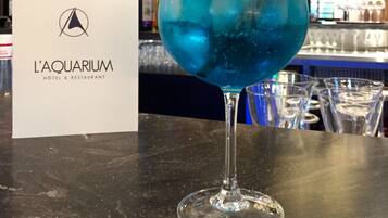 Een cocktailglas met een blauwe drank op een barkruk, een bordje voor L'AQUARIUM HOTEL & RESTAURANT, en planken met diverse flessen op de achtergrond.