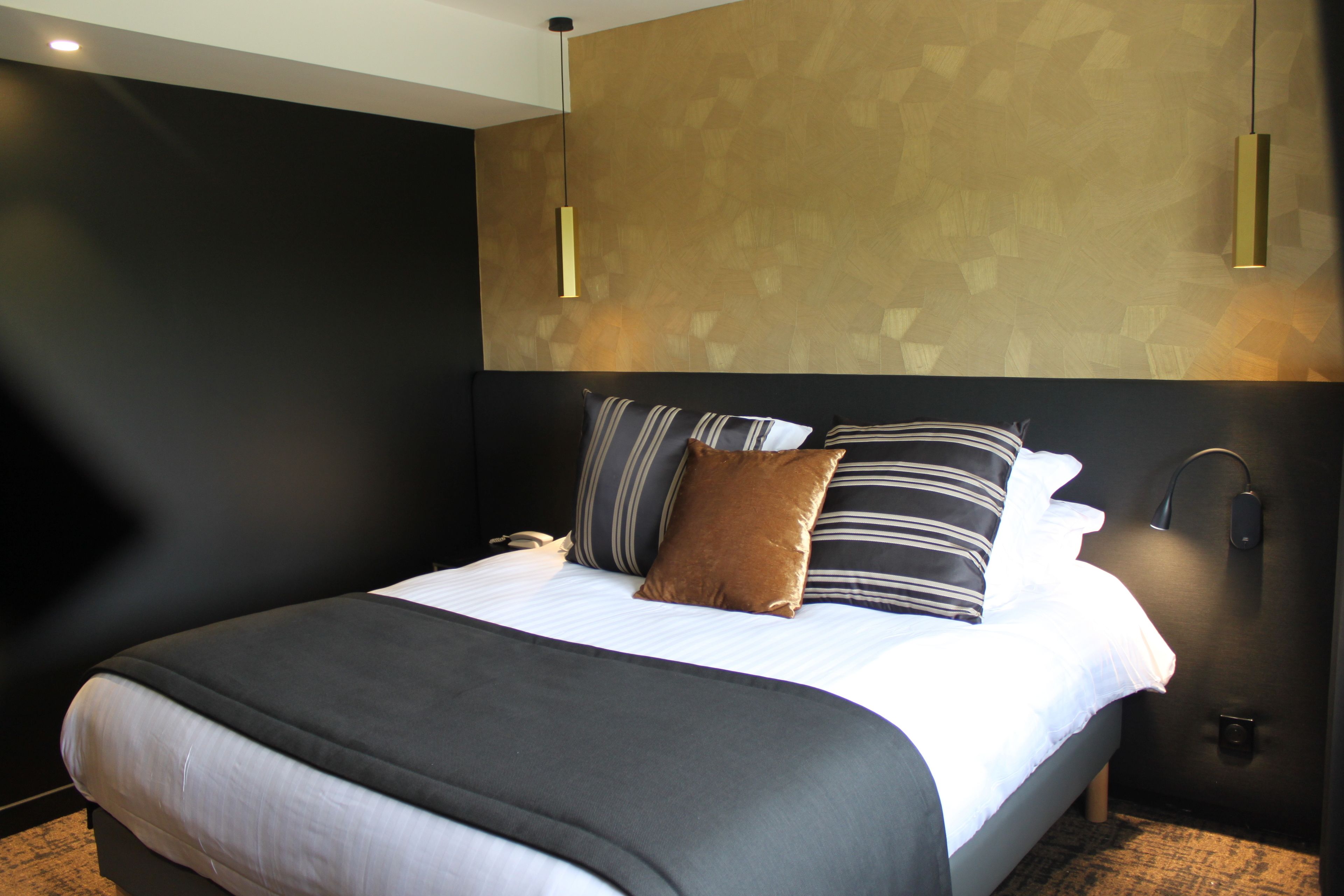 A modern hotel room with a large bed, a headboard with a geometric pattern, and a wall-mounted light fixture.