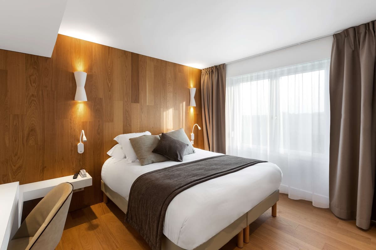 Ein modernes Hotelzimmer mit einer Wandverkleidung aus Holz, einem großen Bett mit grauer Decke, zwei Nachttischlampen, einem Nachttisch mit Telefon und einem Fenster mit Vorhängen.