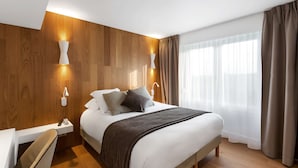 Una habitación de hotel moderna con un panel de pared de madera, una cama grande con una manta gris, dos lámparas de noche, una mesita de noche con un teléfono y una ventana con cortinas.