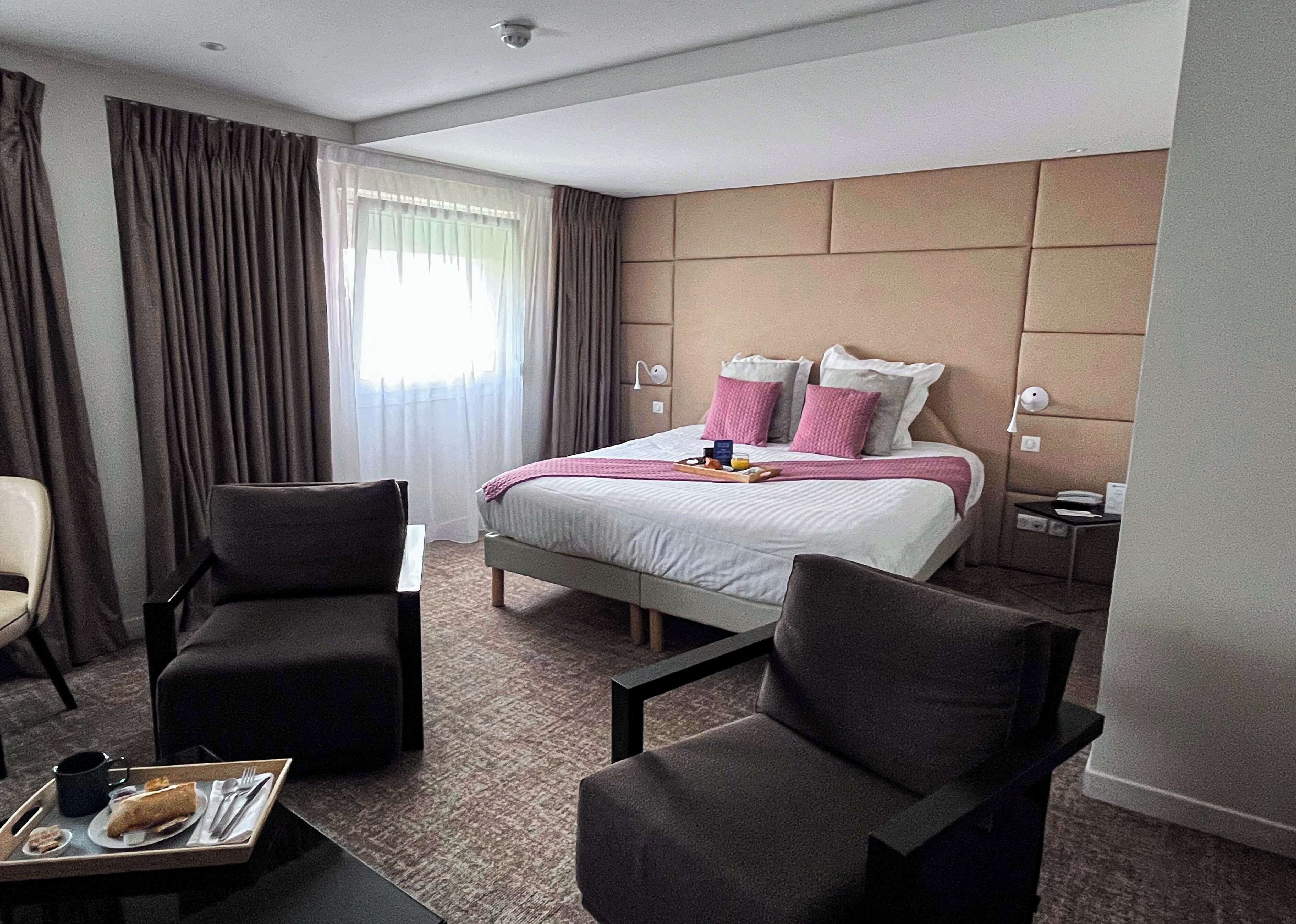 A hotel room with a bed, two armchairs, a small table with a tray of food, and a nightstand with a lamp.