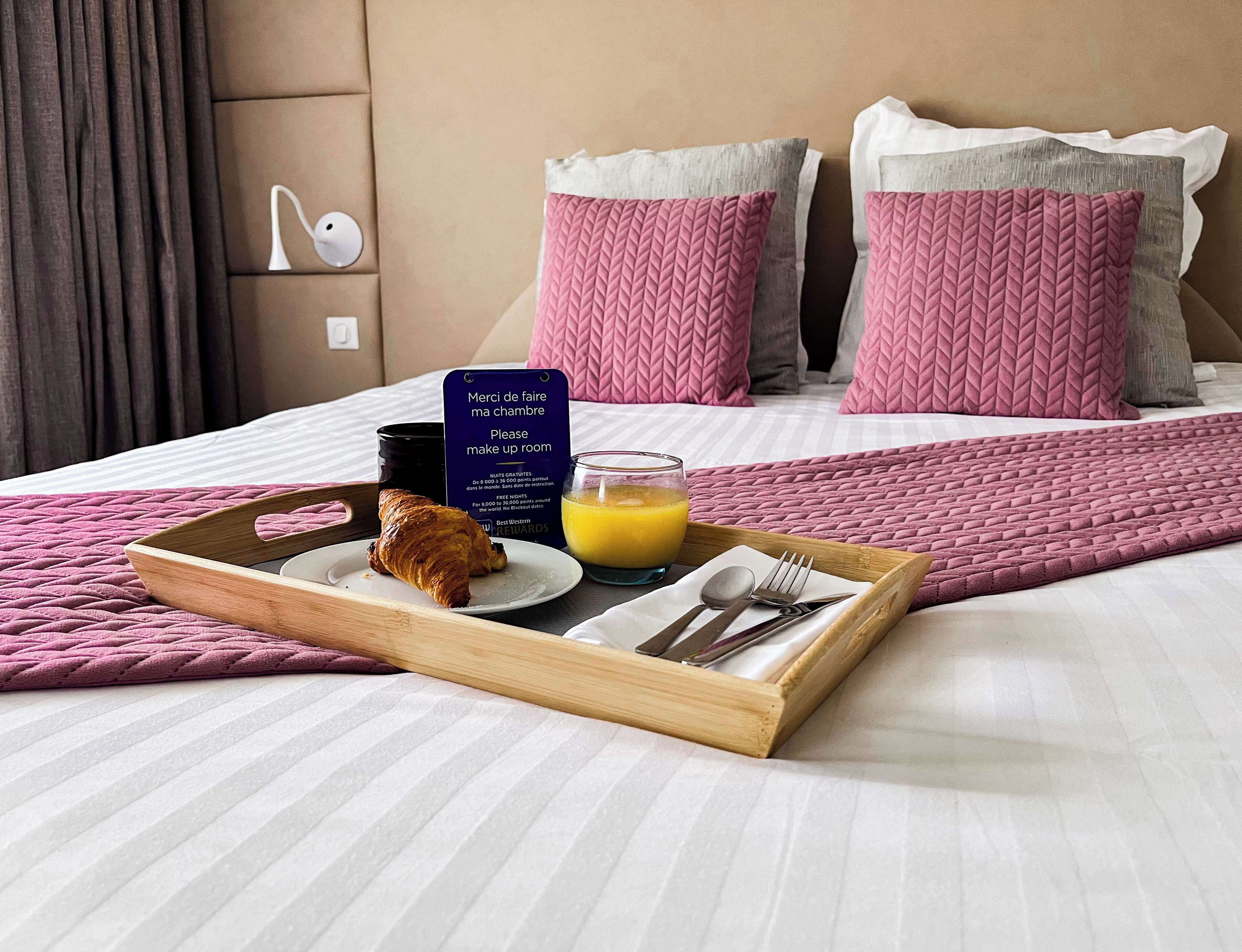 A neatly made-up hotel room with a bed, pink and grey pillows, a breakfast tray with croissant, orange juice, and breakfast utensils.