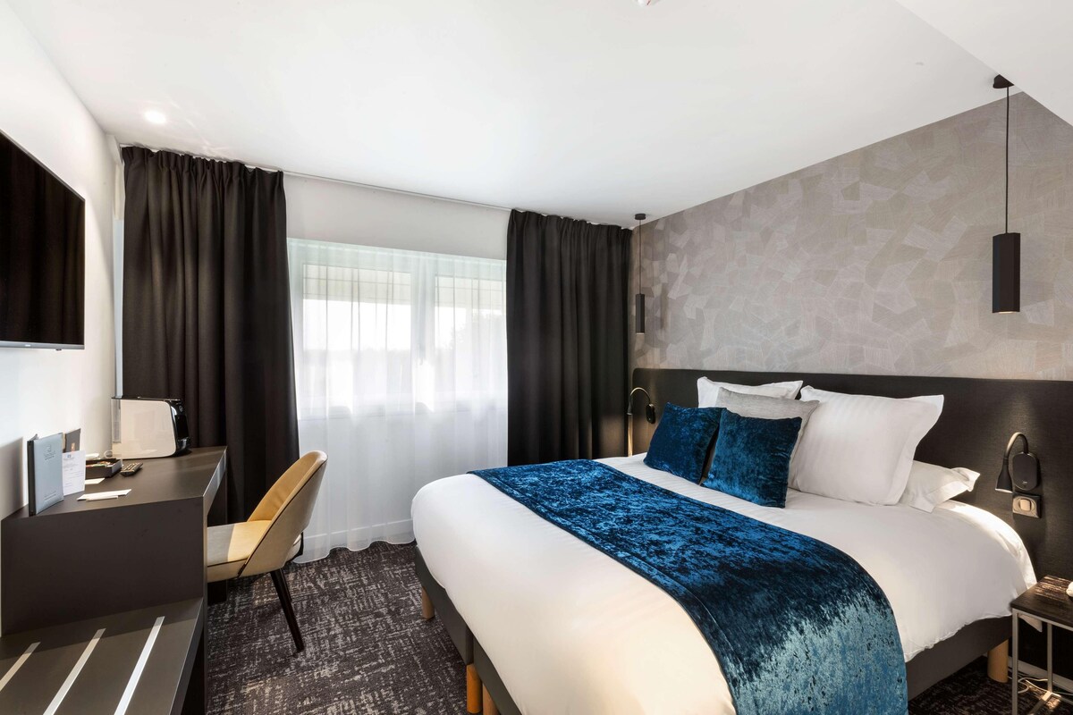 Una habitación de hotel moderna con una cama grande, un escritorio y una silla. Hay una ventana con cortinas, una lámpara de pared y un televisor de pantalla plana.