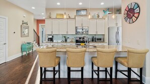 Una cocina moderna con armarios blancos, una isla central con taburetes, electrodomésticos de acero inoxidable y elementos decorativos en las paredes.