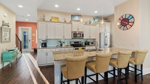 Una cocina moderna con armarios blancos, encimera de granito y electrodomésticos de acero inoxidable. Hay tres taburetes de bar y un reloj grande en la pared.