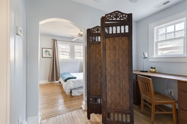 Un dormitorio con una cama, un escritorio y una silla. Se aprecia una ventana con persianas, una puerta y un cuadro en la pared.