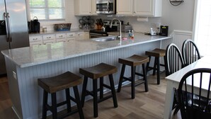 Una cocina con armarios blancos, un refrigerador de acero inoxidable, un microondas y un fregadero. Hay cuatro taburetes de madera y una mesa con sillas.