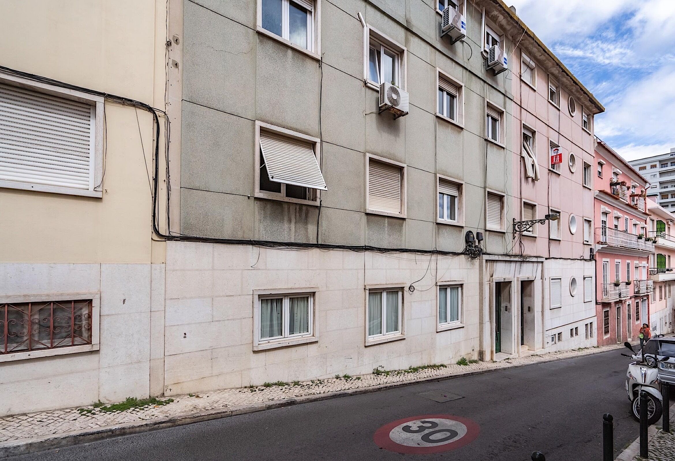 Un immeuble à plusieurs étages, doté de nombreuses fenêtres, dont certaines sont équipées de volets, et un panneau indiquant la limite de vitesse est placé sur la rue.