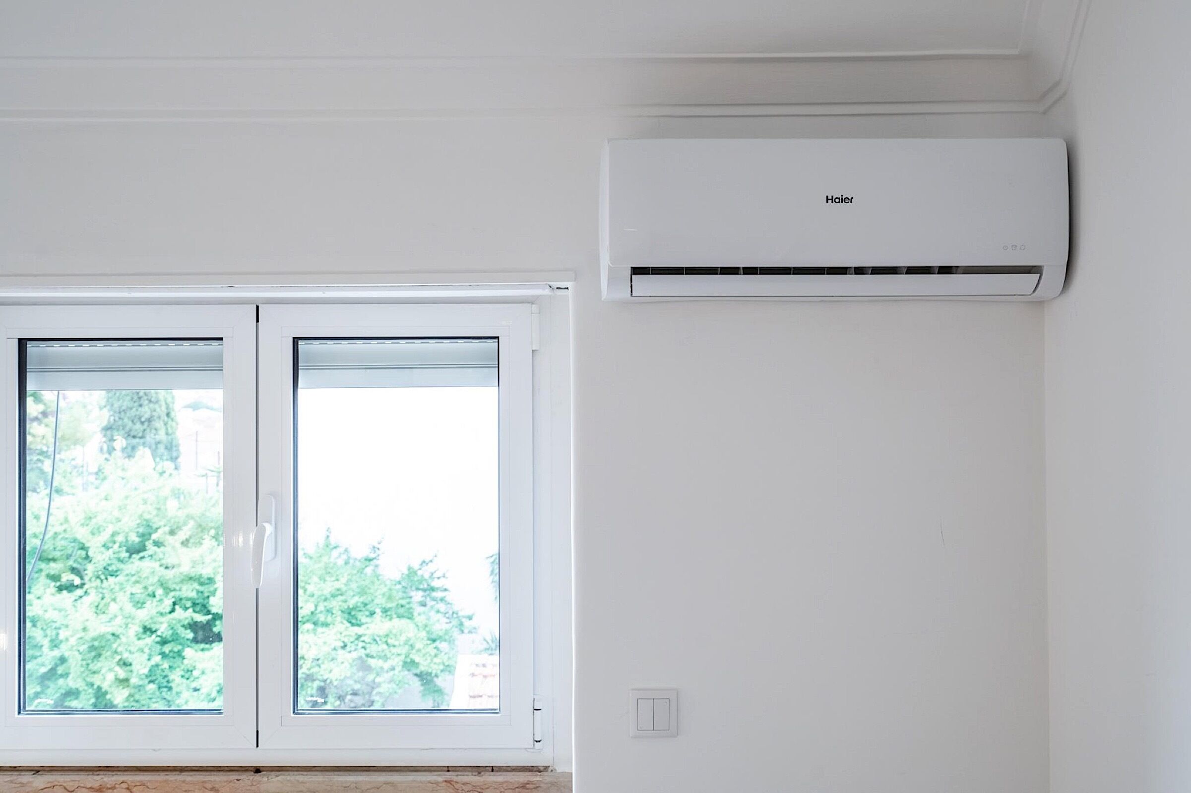A room with a white air conditioning unit mounted on the wall and a window with a view of greenery outside.