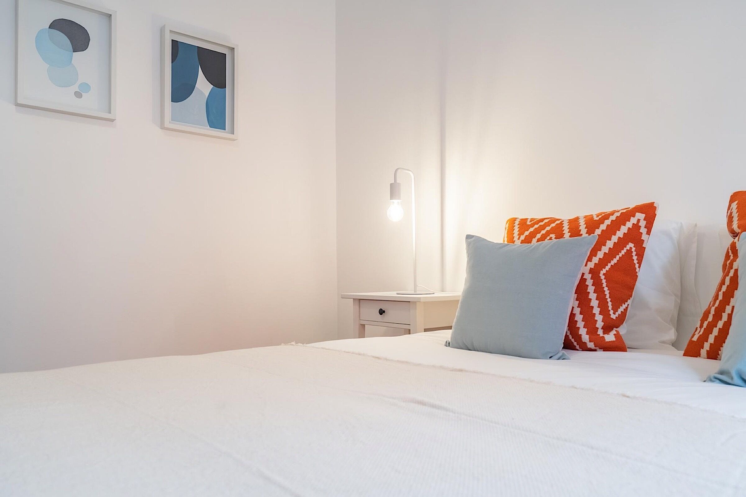 A neatly made bed with white bedding and orange patterned pillows, a bedside table with a lamp, and two abstract art pieces on the wall.