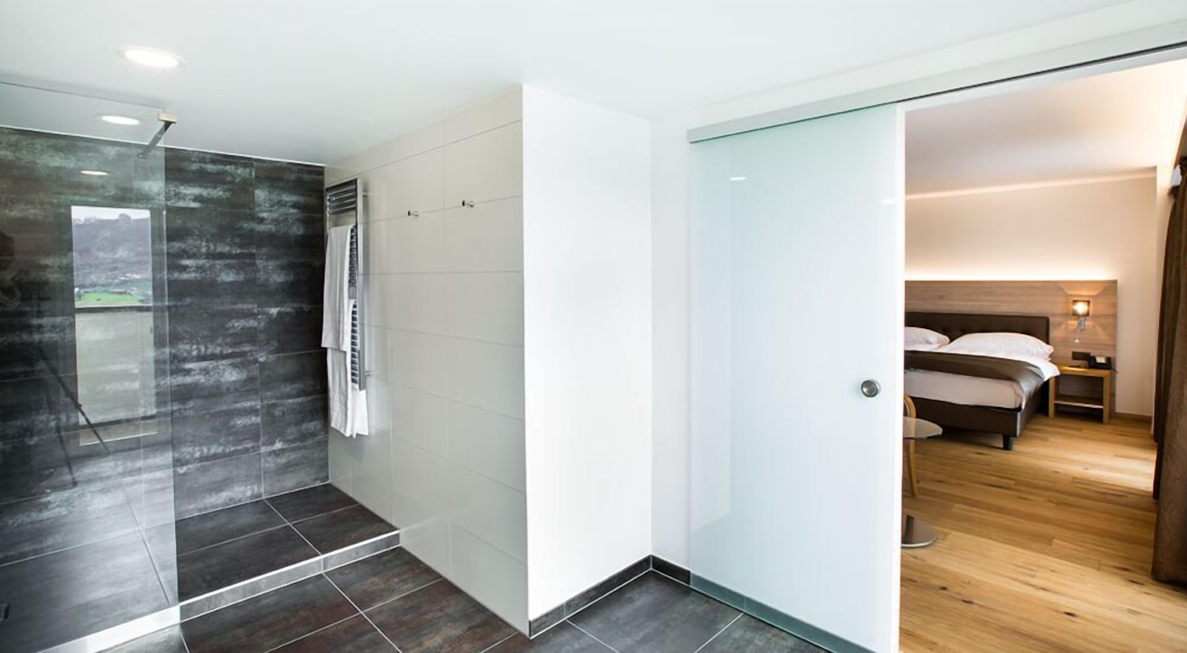 Ein modernes Badezimmer mit Duschkabine aus Glas, weiß gefliestem Duschbereich sowie ein separates Schlafzimmer mit Holzboden, Bett und Nachttisch.