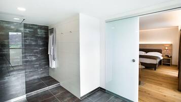Ein modernes Badezimmer mit Duschkabine aus Glas, weiß gefliestem Duschbereich sowie ein separates Schlafzimmer mit Holzboden, Bett und Nachttisch.