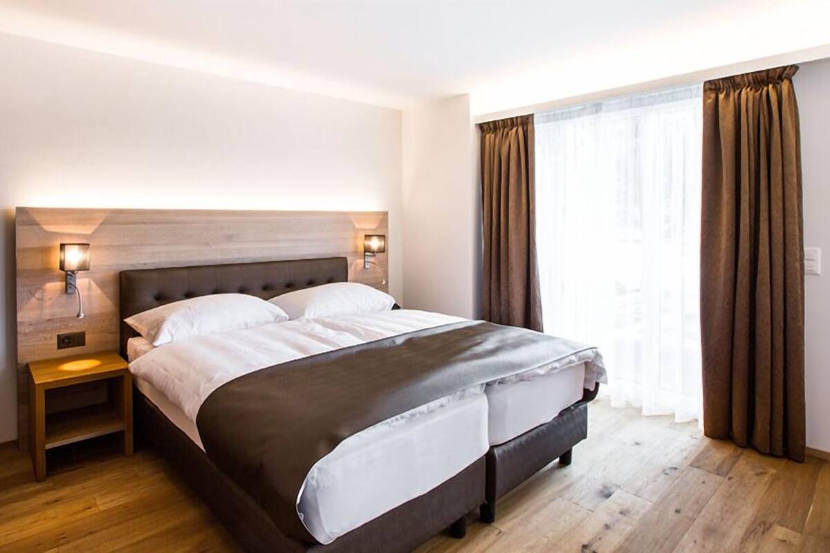 Een moderne hotelkamer met een groot bed, houten vloeren en uitzicht op de stad door grote ramen.