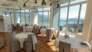 Een restaurant met ronde tafels en stoelen, grote ramen met een prachtig uitzicht op bergen en een meer.