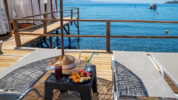 A wooden deck with two lounge chairs, a table with fruit and drinks, and a view of the sea with boats. - Assos Villa Natura (Ayvacik)