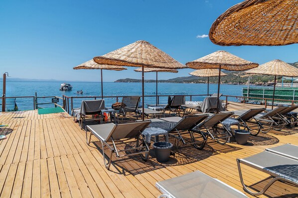 A wooden deck with lounge chairs and thatched umbrellas overlooking a body of water. - Assos Villa Natura (Ayvacik)