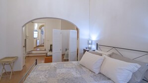 A bedroom with a bed, bedside table, and a lamp. There is a wooden chair and a small table. An arched doorway leads to another room.