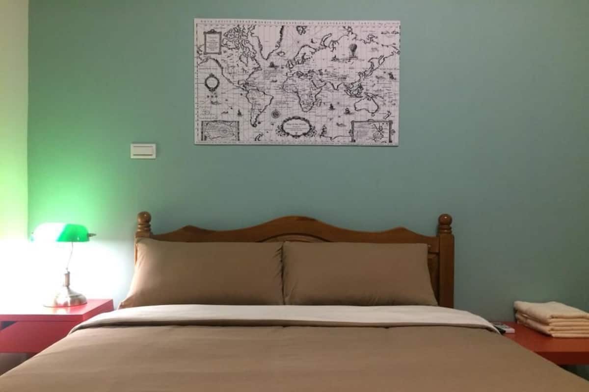Una cama bien hecha con cabecera de madera, un mapa del mundo en la pared y una lámpara verde sobre una mesita de noche.