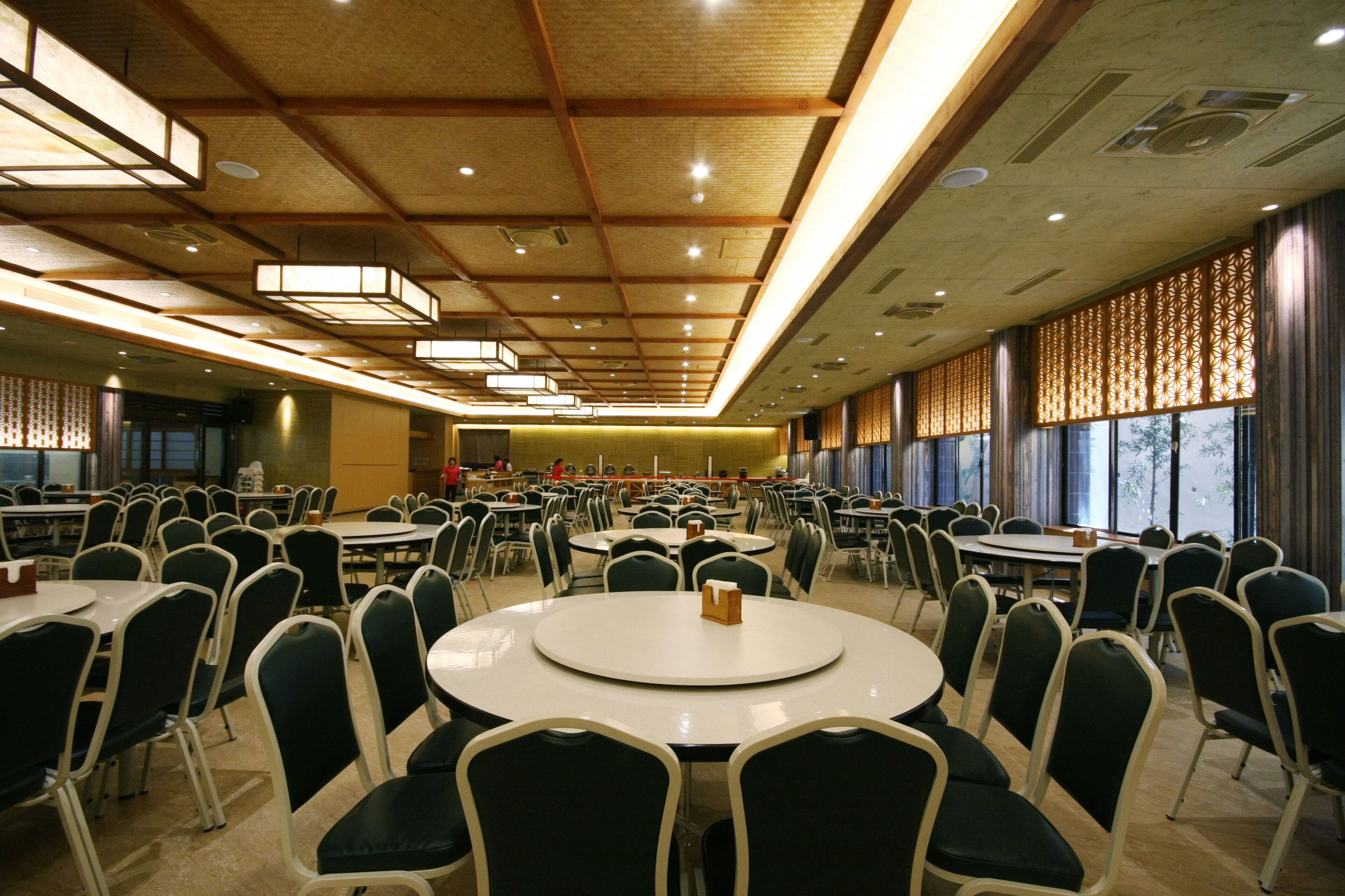 Un comedor amplio con varias mesas redondas y sillas, techos altos con vigas de madera y ventanas grandes con cortinas decorativas.