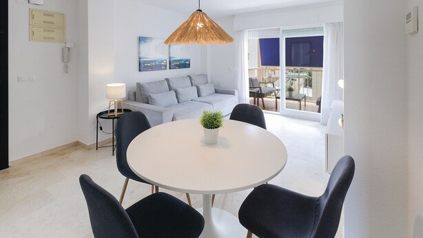 Ein modernes Wohnzimmer mit einem runden weißen Tisch, dunkelblauen Stühlen, einem grauen Sofa und einer Pendelleuchte.