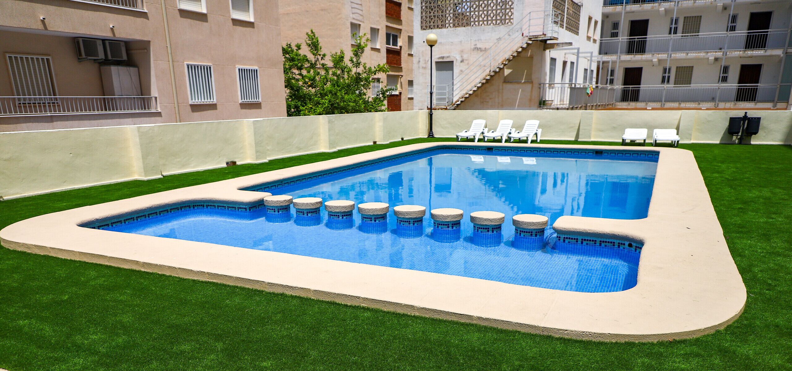 A rectangular pool with a tiled edge and a surrounding grassy area. There are lounge chairs and a staircase leading to an upper level.