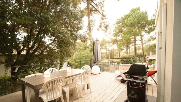Patio esterno con tavolo e sedie in legno, barbecue e ringhiera in vetro che si affaccia su un giardino.