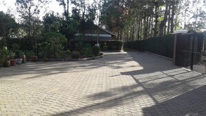 Un camino pavimentado que conduce a un edificio con acceso cubierto, rodeado de árboles y plantas en macetas.