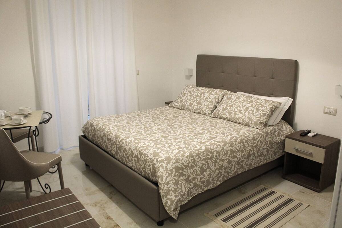A bedroom with a bed, a bedside table, a chair, and a small desk with a cup and saucer.