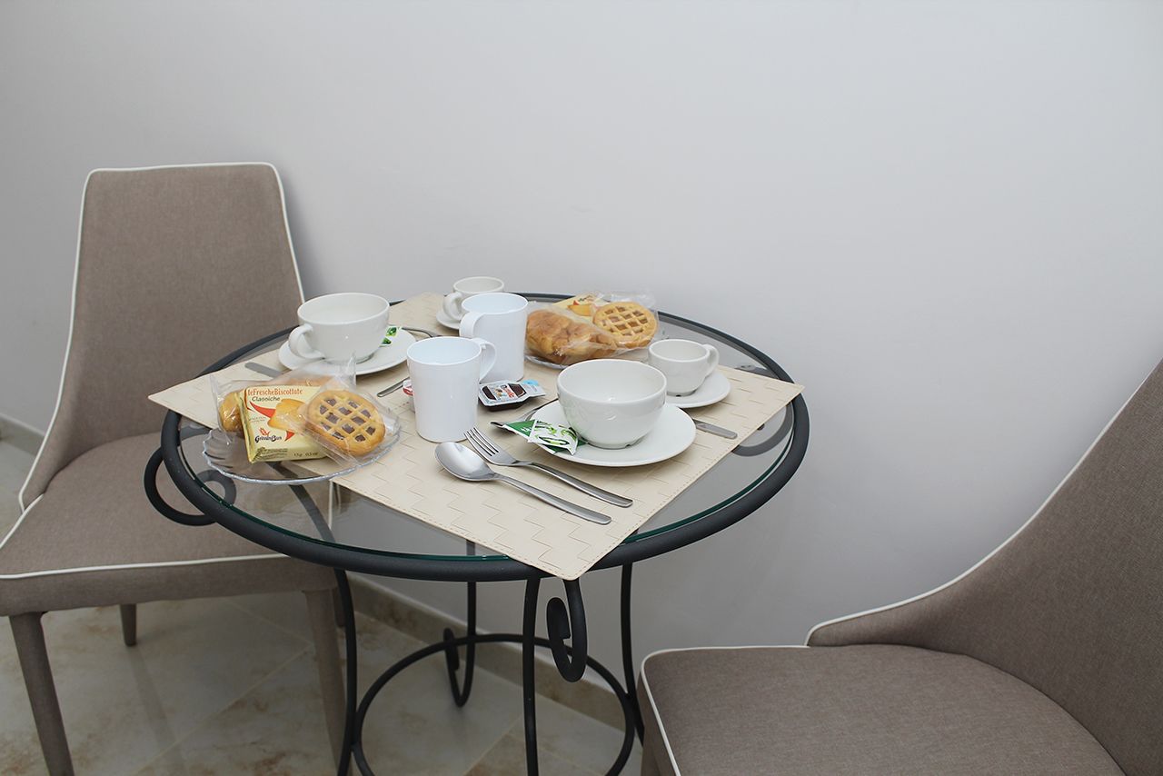 A round glass table with a beige placemat, surrounded by two beige chairs. On the table, there are white cups, a white bowl, a white plate, a white teapot, a white sugar bowl, a white fork, a white spoon, a white napkin, and a package of cookies.