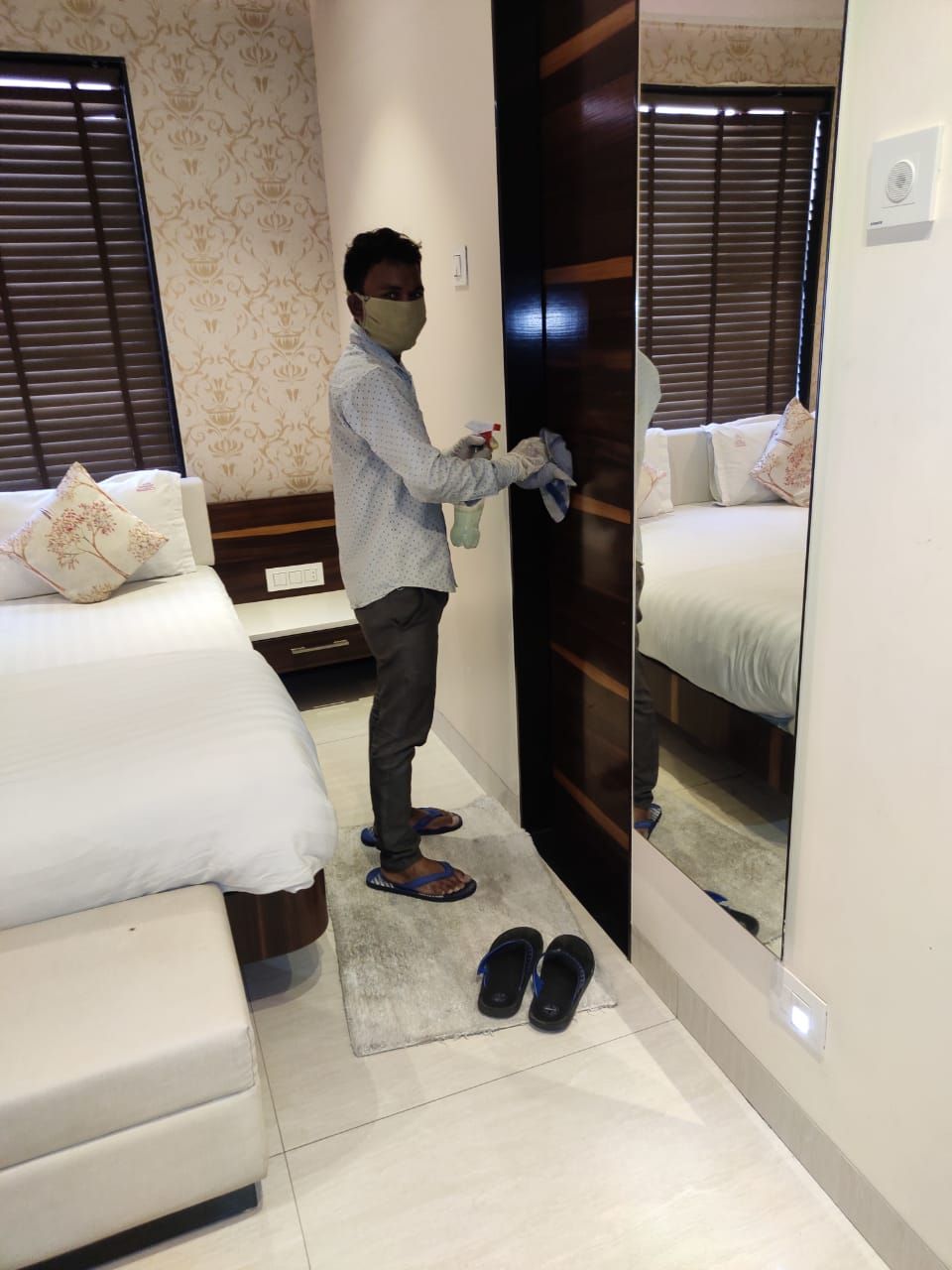 A hotel room with a bed, a mirror, and a pair of slippers on the floor.