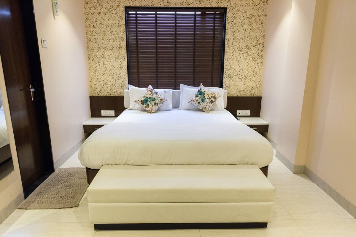 A hotel room with a large bed, a bench, a wall-mounted control panel, and a window with blinds.