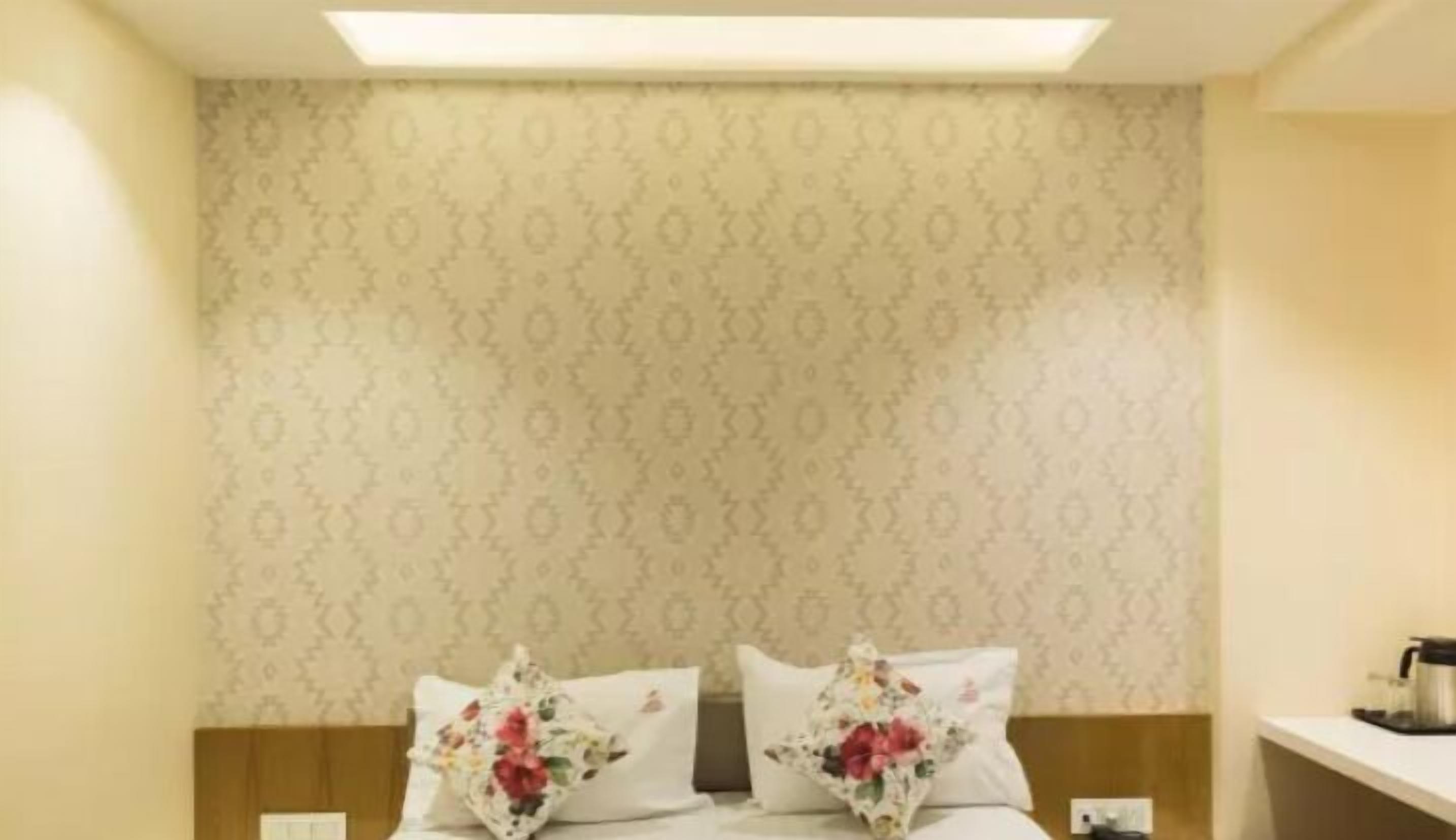 A hotel room with a patterned wallpaper, a bed with floral pillows, and a small table with a kettle.