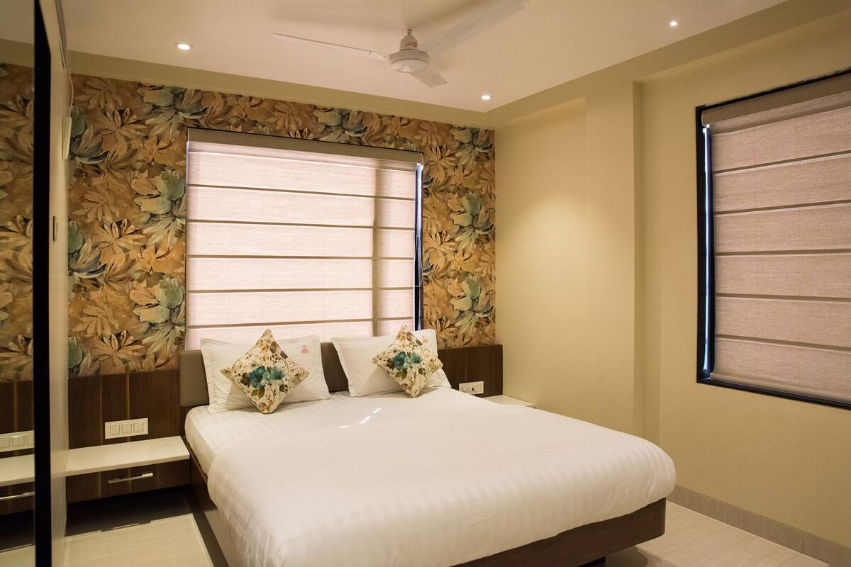 A bedroom with a large bed, a ceiling fan, a window with blinds, and a wall with floral wallpaper.