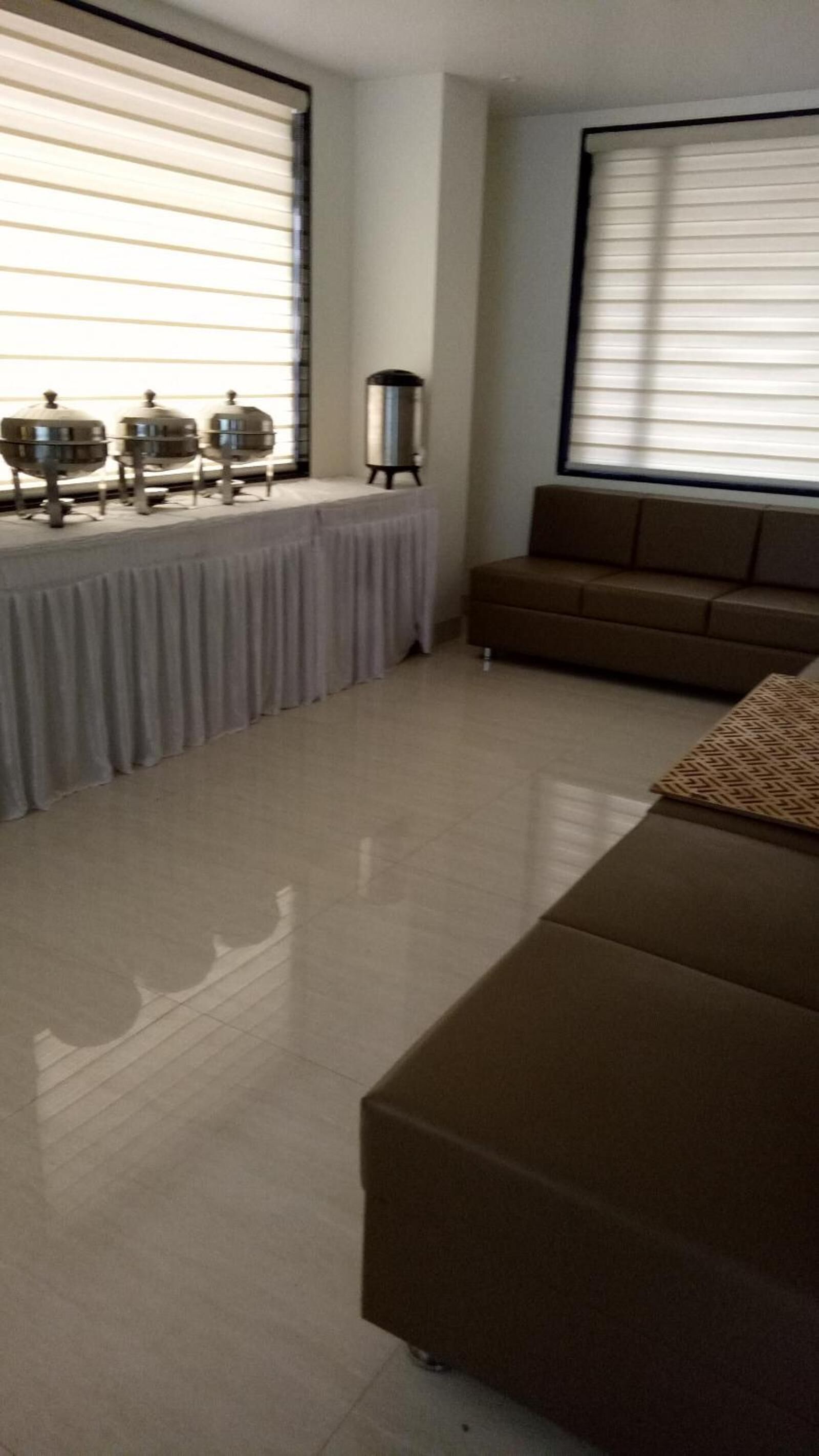A living room with a brown sofa, a coffee table, and a side table with a lamp. There are large windows with blinds, and a radiator on the wall.
