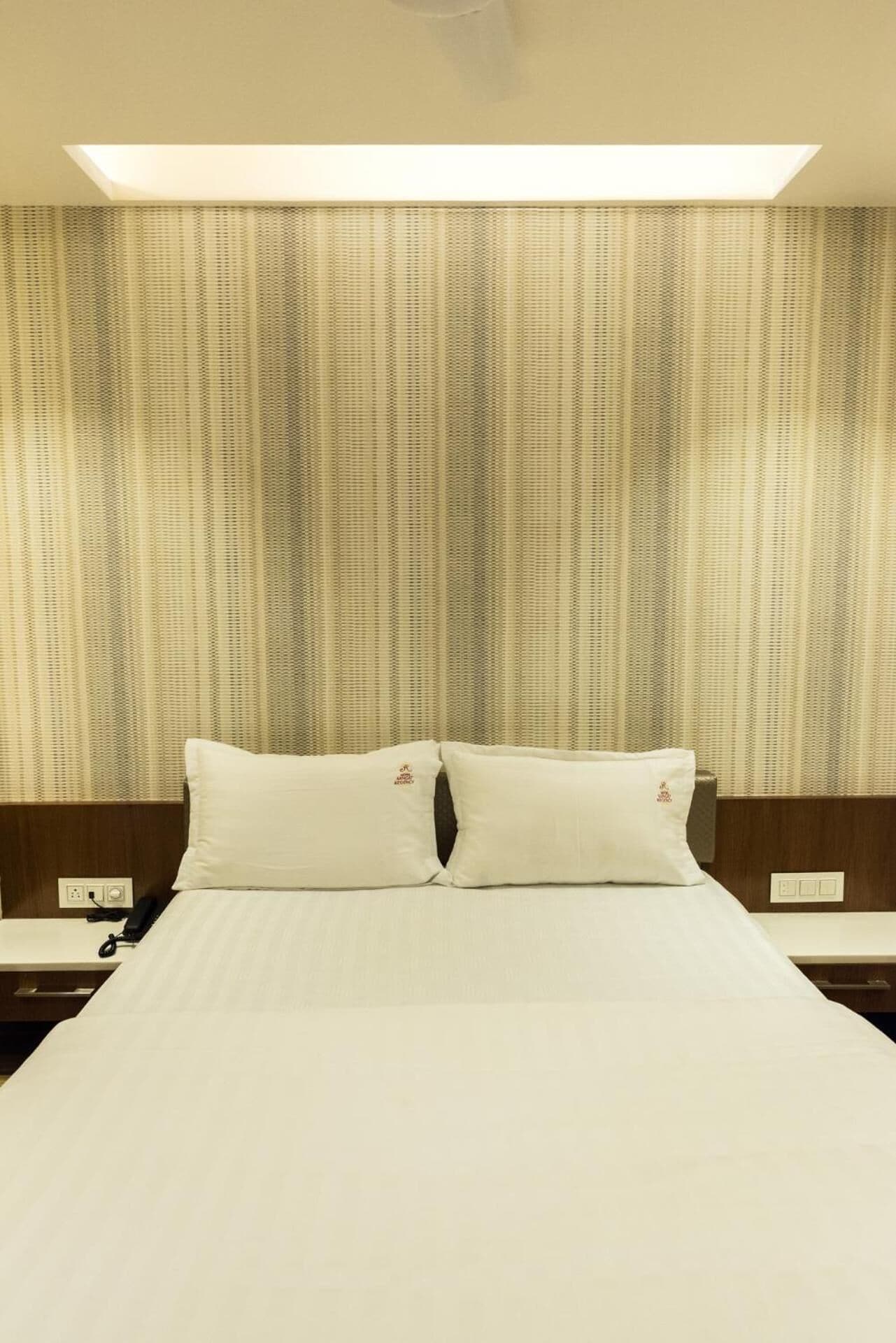 A hotel room with a large bed, two pillows, a nightstand with a phone, and a headboard with a light fixture above.